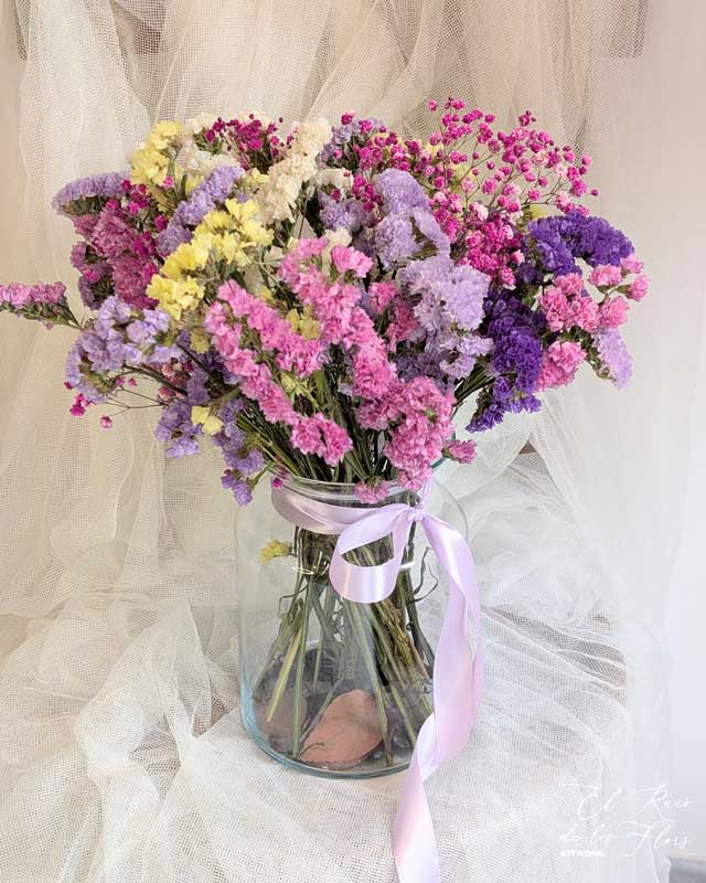 Ramo de statice seco mix de color, con paniculata fucsia seca. Jarrón de cristal con hojas de eucalipto en la base y lazada de raso lila. Medidas aprox: altura jarrón 20,50cm x 14cm diámetro en la base. Ramo diámetro 30cm x 40cm altura total