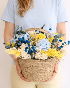 Cesto de mimbre decorado con una alegre combinación de azul y amarillo y blanco. Materiales utilizados: Broom amarillo, broom blanco, cardo blanco, botao azul, Hortensia azul celeste, eucalipto preservado gris.