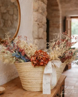 Cesto de mimbre decorado con flores secas en tonos otoñales. Materiales utilizados: Hortensia natural seca, avena salvaje, limonium rojo, limonium benlat lila grisáceo, eucalipto gris preservado. Medidas aprox.: base del cesto 22 cm largo x 11,50 cm profundidad. Con el trabajo floral, altura total 52 cm 70 cm largo